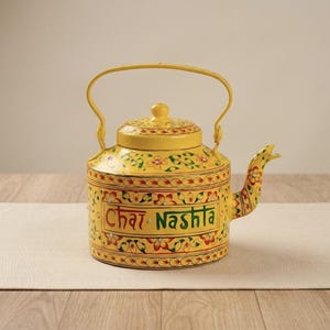 May include: A bright yellow metal teapot featuring a hand-painted floral pattern in red, green, and orange. The front of the teapot displays the text "Chai Nashta" in bold, colorful lettering. It includes a matching lid and a curved handle.