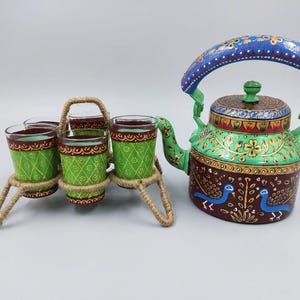 Puede incluir: Una tetera de metal pintada a mano y cuatro vasos a juego en un soporte de cuerda de yute. La tetera tiene un asa azul, cuerpo verde con motivos florales y base roja oscura con pájaros azules. Los vasos presentan patrones de color verde.