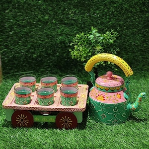 May include: A decorative tea set with a hand-painted kettle and six glasses on a cart. The kettle is green and pink with a yellow handle. The glasses are green and red, and the cart is green with red wheels. The set is placed on a green surface.