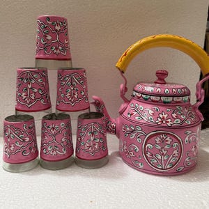 Op de afbeelding: Een roze metalen theepot met een geel handvat en een bijpassende set van zes roze glazen. Zowel de theepot als de glazen zijn voorzien van handgeschilderde witte, groene en rode bloemmotieven met gedetailleerde ranken op de oppervlakken.