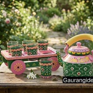 May include: A hand-painted metal tea set featuring a pink and green kettle with a yellow handle, accompanied by a matching green cart on wheels holding four small glass tumblers. The set displays floral patterns and the text Gaurangidecor.