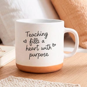 Puede incluir: Una taza de cerámica blanca con acabado moteado y base de color terracota. El frente presenta texto negro en cursiva que dice: "Teaching fills a heart with purpose", acompañado de dos pequeños iconos de corazón a los lados del texto.