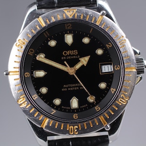 Może przedstawiać: Srebrno-złoty zegarek Oris z czarną tarczą. Zegarek posiada złote wskazówki i znaczniki, okienko daty oraz czarny skórzany pasek. Jest to zegarek automatyczny, wodoszczelny do 100 metrów.