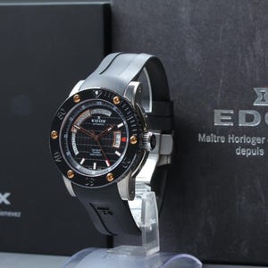 May include: An Edox automatic wristwatch with a black dial, silver-tone hands, and a black rubber strap. The watch features a rotating bezel with gold-tone accents, a day-date display, and the text Edox Maître Horloger - Les Genevez depuis 1884.