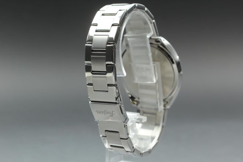May include: A silver-toned wristwatch with a metal band. The watch is displayed on a clear stand, showcasing the band's links and the watch face. The word "fugon" is visible on the band.