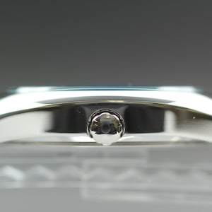 May include: A side view of a silver wristwatch. The watch has a rectangular face with a silver case and a small, round crown. The watch band is also silver. The watch is displayed on a clear surface.