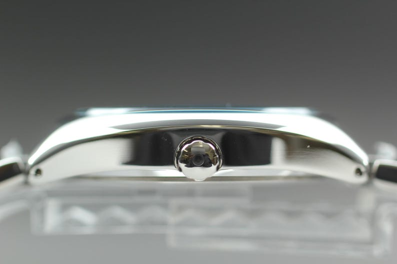 May include: A side view of a silver wristwatch. The watch has a rectangular face with a silver case and a small, round crown. The watch band is also silver. The watch is displayed on a clear surface.
