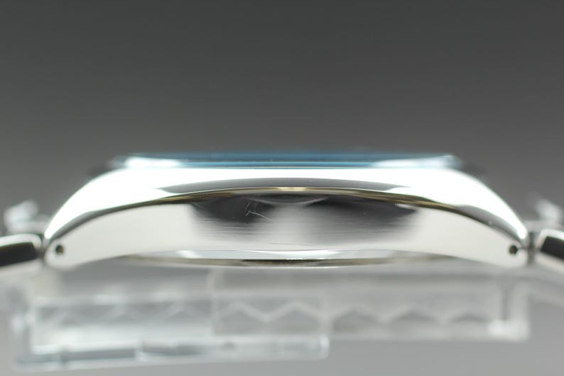 May include: Side view of a silver wristwatch with a light blue face. The watch has a curved, polished metal case and a matching metal band. The watch is displayed against a blurred background.