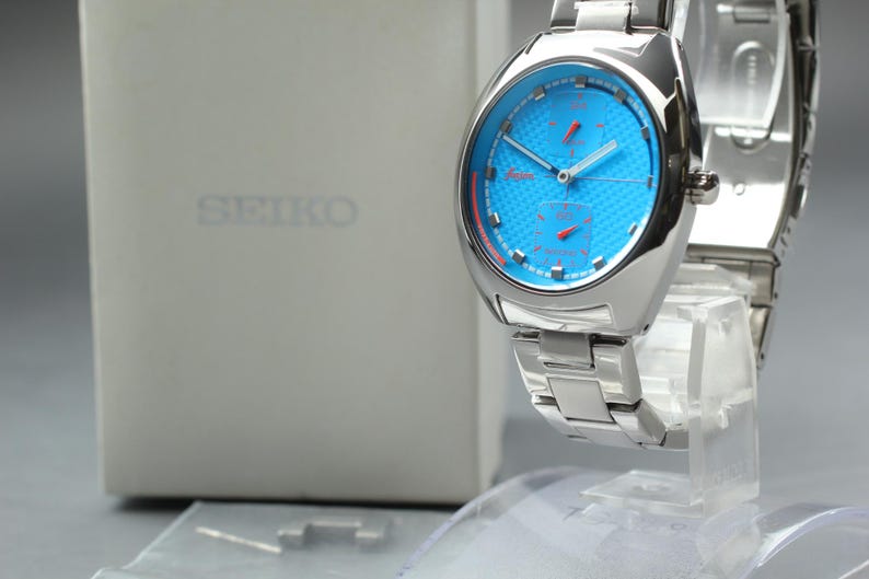 May include: A silver-toned Seiko watch with a vibrant blue face and red accents. The watch features a textured dial and a stainless steel band. The watch is displayed on a clear stand, with the Seiko box in the background.