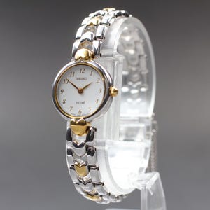May include: A Seiko watch with a silver and gold-tone strap and a white face. The watch has gold-tone hands and hour markers. The watch is displayed on a clear stand. The watch face reads "SEIKO" and "TIME".