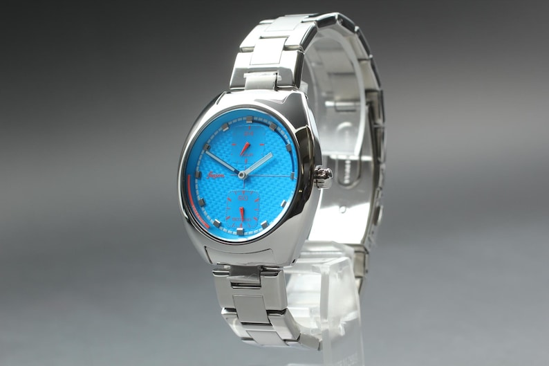 May include: A silver-toned wristwatch with a vibrant blue face and red accents. The watch features a stainless steel band and a unique, modern design. The watch is displayed on a clear stand.