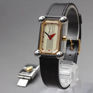May include: A rectangular wristwatch with a black leather band. The watch face is cream-colored with black Roman numerals and a red arrow-shaped hand. The watch has a silver-colored case with gold accents and silver ball-shaped details.