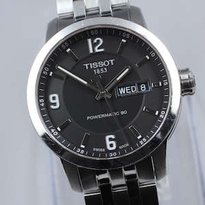 May include: A close-up of a Tissot Powermatic 80 wristwatch with a black dial and silver-tone stainless steel band. The watch face displays the day and date, with the day reading "WED 8". The watch has a silver bezel and a silver band.