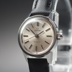 Pode incluir: Um relógio de pulso Omega Genève com um mostrador redondo em tom prateado, marcadores de hora e ponteiros pretos. O relógio possui uma caixa de aço inoxidável polido e uma pulseira de couro preto texturizado com fecho de fivela.