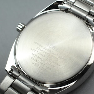 May include: Close-up of the back of a silver ALBA wristwatch. The watch has a stainless steel band and a round, brushed metal back with engraved text. The text includes "WATER RESISTANT SHAR", "STAINLESS STEEL", and "MOVEMENT JAPAN".