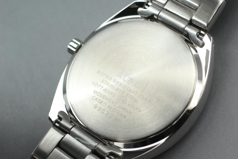 May include: Close-up of the back of a silver ALBA wristwatch. The watch has a stainless steel band and a round, brushed metal back with engraved text. The text includes "WATER RESISTANT SHAR", "STAINLESS STEEL", and "MOVEMENT JAPAN".