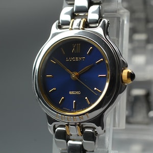 Könnte beinhalten: Silber-goldfarbene Seiko Lucent Armbanduhr mit tiefblauem Zifferblatt. Die Uhr hat goldene Zeiger und Markierungen, mit dem Text "LUCENT" und "SEIKO" auf dem Zifferblatt. Das Armband ist silberfarben mit goldenen Akzenten.