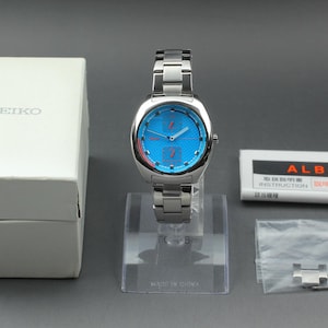 May include: A silver-toned Alba watch with a blue face and matching stainless steel band. The watch is displayed on a clear stand, with a white Seiko box and Alba instruction booklet nearby. A small bag with extra links is also present.