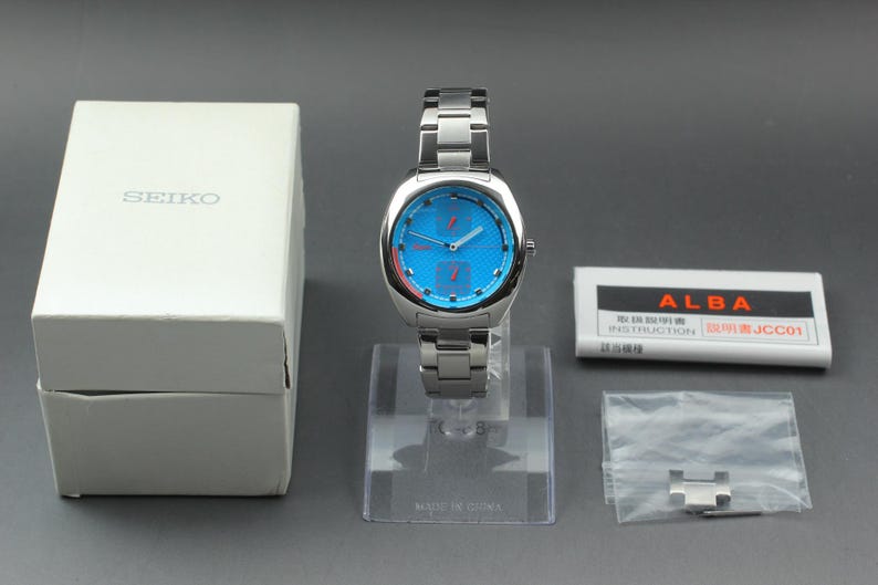 May include: A silver-toned Alba watch with a blue face and matching stainless steel band. The watch is displayed on a clear stand, with a white Seiko box and Alba instruction booklet nearby. A small bag with extra links is also present.