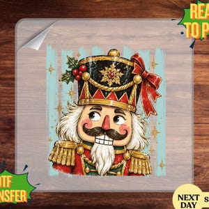 May include: A DTF transfer featuring a nutcracker illustration with a black hat, red bow, and gold accents. The nutcracker has a white beard, mustache, and rosy cheeks, set against a light blue background with gold star accents and holly.