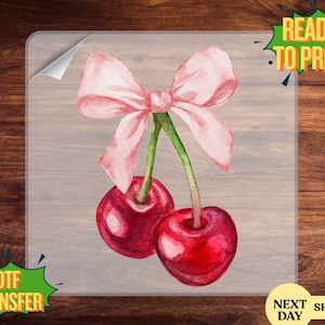 May include: A watercolor design featuring two ripe cherries with a pink bow and green stems. The image is labeled "READY TO PRESS" and "DTF TRANSFER". The design is on a transparent sheet.