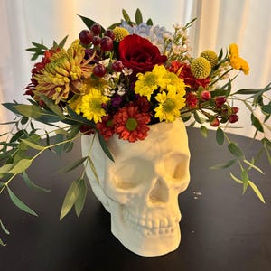 Pode incluir: Um vaso branco em forma de caveira de cerâmica preenchido com um arranjo floral vibrante. As flores incluem rosas vermelhas, margaridas amarelas e folhagem verde. O vaso está sobre uma superfície preta.