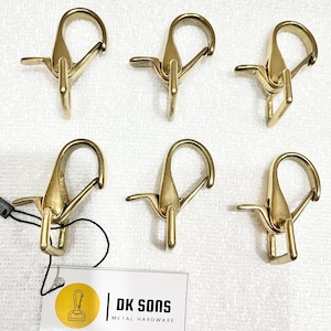 Puede incluir: Seis cierres giratorios de metal color latón. Cada cierre tiene una puerta con resorte y una base giratoria. Los cierres están dispuestos sobre una superficie blanca. Una etiqueta con el texto "DK SONS METAL HARDWARE" está adjunta.