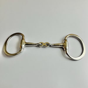 May include: A silver-colored horse bit with a central joint and rounded rings on each end. The bit is designed for use in equestrian activities and is made of a shiny metal.