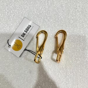 May include: Two gold-colored metal hardware clasps with a hook design. A white tag with the text "DK SONS METAL HARDWARE" is attached to one clasp. The clasps are on a white, textured surface.