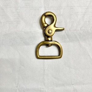 Puede incluir: Un cierre giratorio de color latón con una base en forma de D y un gancho con resorte. El cierre está diseñado para sujetarse a bolsos, correas u otros accesorios. El metal tiene un aspecto ligeramente envejecido.