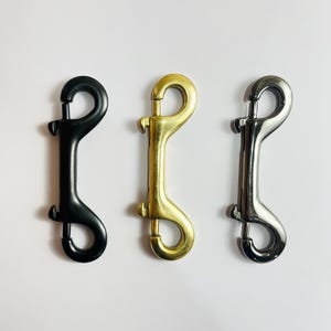 Puede incluir: Tres mosquetones de resorte metálicos en negro, dorado y plateado. Los ganchos están diseñados con un ojal giratorio y una puerta con resorte para una fijación segura. Los mosquetones se muestran sobre un fondo blanco.