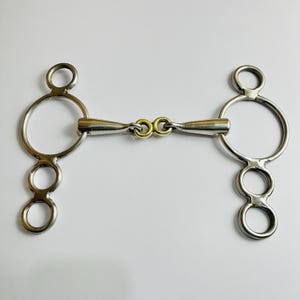 May include: A stainless steel horse bit with a gold-colored center link. The bit features large circular rings and smaller rings below. The metal has a polished finish, reflecting light. This equestrian equipment is designed for use with a bridle.