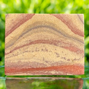 Peut inclure: Une pierre rectangulaire avec un motif en couches de teintes beige, rouge et grise. La surface de la pierre présente un aspect naturel et texturé, avec des bandes ondulées de couleurs sur la face. L'esthétique générale est terreuse et organique.