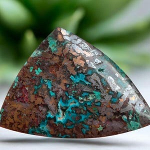 May include: A triangular-shaped gemstone with a mix of colors. The stone features a blend of brown, teal, silver, and red hues. The surface has a textured appearance, with a polished finish. The gemstone is likely used for jewelry or decorative purposes.