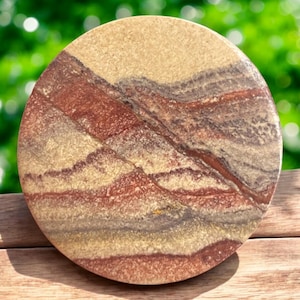 May include: A round, natural stone coaster with a layered pattern of tan, red, and gray hues. The coaster has a smooth surface and a polished finish. The stone is set against a blurred green background and a wooden surface.