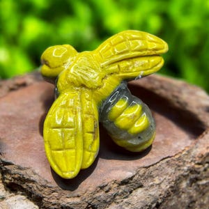 May include: A carved yellow bee figurine with detailed wings and body segments. The bee has a gray and yellow abdomen. The figurine is set against a natural, textured background, with a blurred green backdrop.
