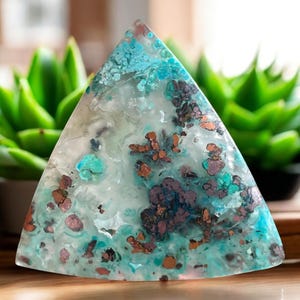 May include: A triangular-shaped decorative stone with a unique pattern. The stone features a mix of turquoise, brown, and white colors. The stone is set against a blurred background of green plants.