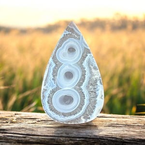 Può includere: Una fetta di agata lucidata a forma di goccia con tre distinte bande circolari concentriche di colore bianco e grigio traslucido. La pietra presenta una consistenza cristallina lungo i bordi esterni e una superficie liscia e lucida.