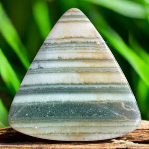 Può includere: Una pietra triangolare lucidata con bande orizzontali di colore. La pietra presenta strati di bianco, beige e sfumature di verde. La pietra è liscia e ha bordi arrotondati. Lo sfondo è un fogliame verde sfocato.