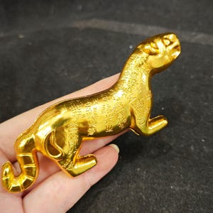May include: A metallic gold-coloured figurine shaped like a crouching tiger. The surface of the tiger is covered in intricate, embossed Chinese characters throughout its body, from the head down to the curved tail, with a smooth, reflective finish.