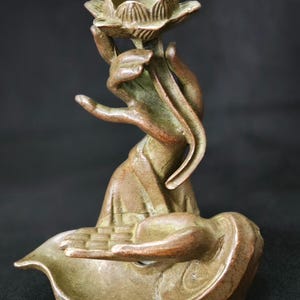 Puede incluir: Escultura de bronce de una mano sosteniendo una flor de loto. La escultura tiene una apariencia envejecida y antigua con tonos verdes y marrones. La mano está elegantemente posada y la flor de loto está intrincadamente detallada.