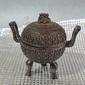May include: A silver-colored, spherical incense burner with a detailed, openwork design. It features two handles, a decorative lid with a small sculpted figure, and three short legs. The burner has an antique aesthetic.