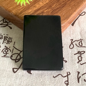 May include: A rectangular, solid black object with rounded corners. The object has a glossy, reflective surface. The item is displayed on a patterned, beige fabric and a wooden surface.