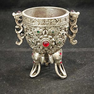 Könnte beinhalten: Ein silberfarbenes Metallgefäß mit detaillierten floralen Reliefmustern, zwei verzierten Seitengriffen und drei elefantenförmigen Beinen. Die Außenseite ist mit eingebetteten roten und grünen Steinen sowie einem Band chinesischer Schriftzeichen verziert.