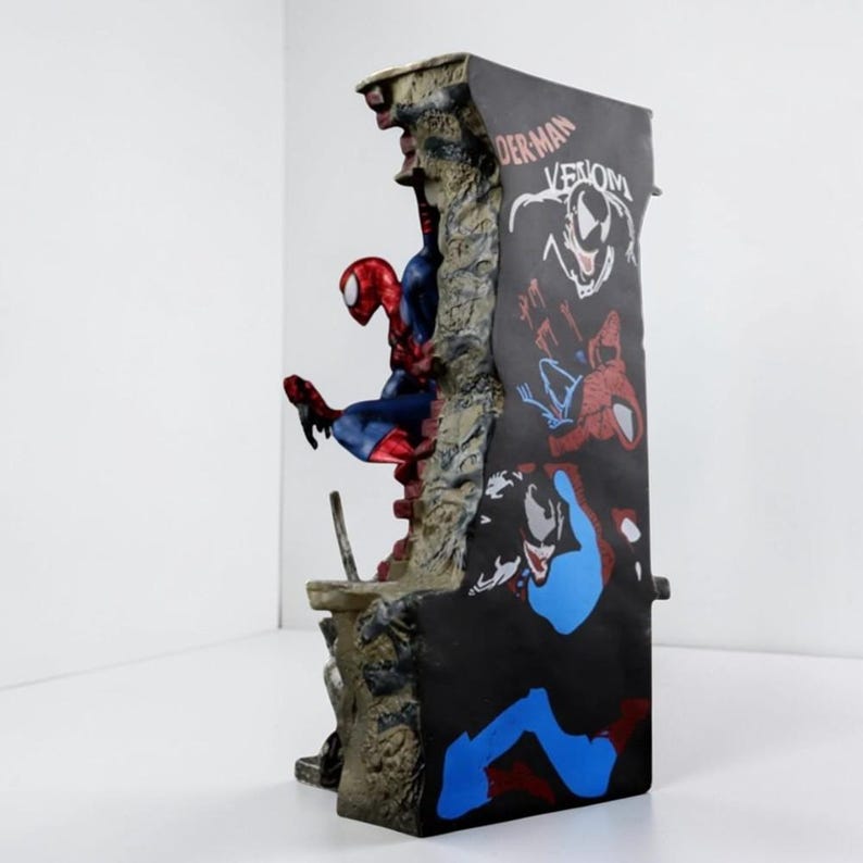 Puede incluir: Figura detallada de Spider-Man en una pose din&aacute;mica, sobre un fondo con Venom y otros gr&aacute;ficos de Spider-Man. La figura es predominantemente roja y azul, con una base texturizada y desgastada.