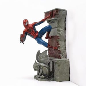 Puede incluir: Figura de Spider-Man trepando una pared de ladrillos. La figura lleva un traje rojo y azul. Una g&aacute;rgola est&aacute; en la base de la pared. La pared es gris con detalles de ladrillo rojo.