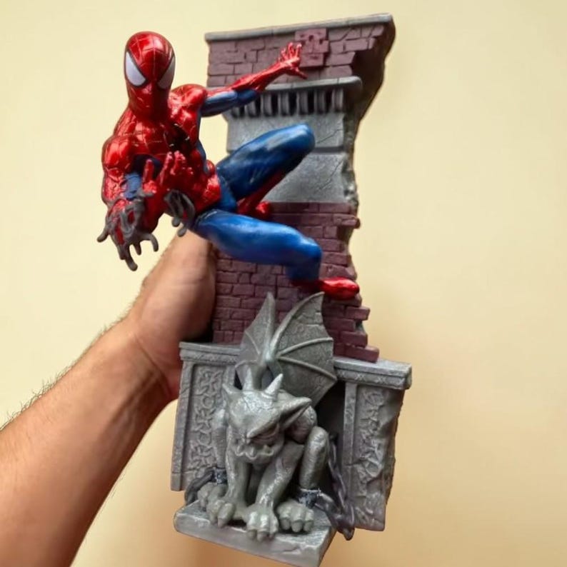 Puede incluir: Una figura de Spider-Man en una pose din&aacute;mica, vestido con un traje rojo y azul, posado en una pared de ladrillo con una g&aacute;rgola. La estatua est&aacute; detallada con un acabado brillante, capturando la imagen del ic&oacute;nico superh&eacute;roe.