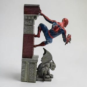 Puede incluir: Una figura de Spider-Man en una pose din&aacute;mica, trepando por una estructura de ladrillo y piedra. La figura lleva un traje cl&aacute;sico rojo y azul. Una estatua de g&aacute;rgola est&aacute; debajo. La estructura tiene un aspecto envejecido.