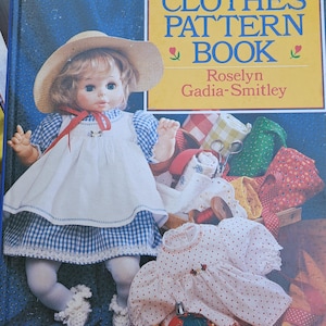 May include: A book titled "Dolls' Clothes Pattern Book" by Roselyn Gadia-Smitley. The cover features a doll wearing a blue and white gingham dress and a white apron. The doll is holding a spool of thread and a pair of scissors.