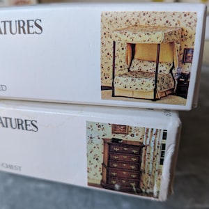 May include: Two white boxes with images of miniature furniture. One box shows a canopy bed with floral patterned fabric. The other box shows a miniature chest of drawers with a floral wallpaper background.